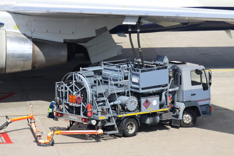 Airliner wing with exposed landing gear and ground support equipment on the tarmac, including hoses and a service truck nearby.