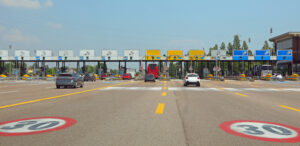 Multi-lane toll plaza with cars approaching booths under a clear blue sky, overhead signs visible above each lane.