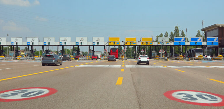 Multi-lane toll plaza with cars approaching booths under a clear blue sky, overhead signs visible above each lane.