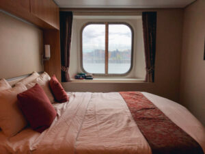 Bed with pink bedding and burgundy cushions in a ship cabin, facing a round window with water and buildings outside