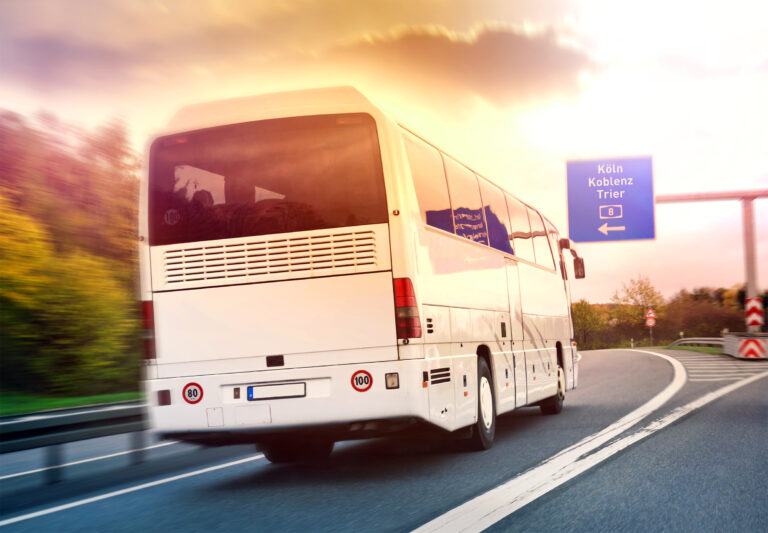 White tour bus driving on a curving highway at sunset, with a blue road sign reading Köln Koblenz Trier ahead.